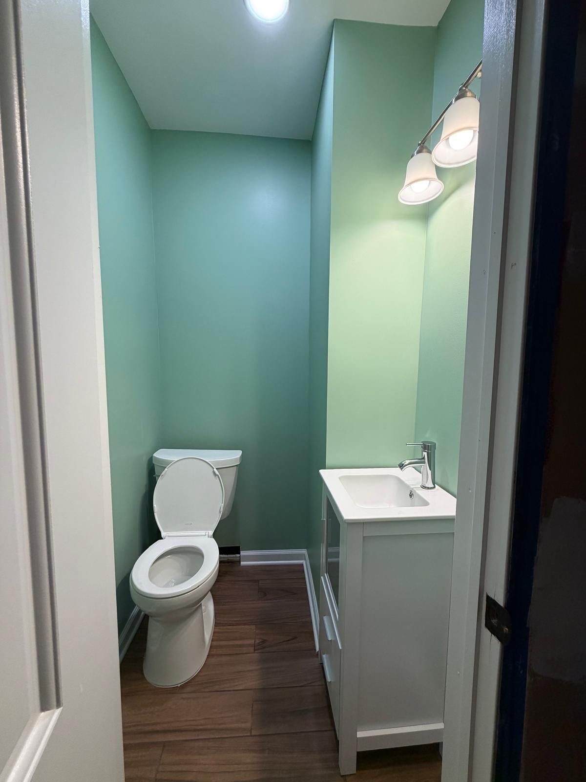 Narrow mint-green bathroom with toilet, white vanity sink, and wall light above dark wood floor.