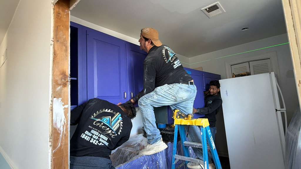 Three construction workers install bright purple kitchen cabinets while standing on a step ladder in a room.