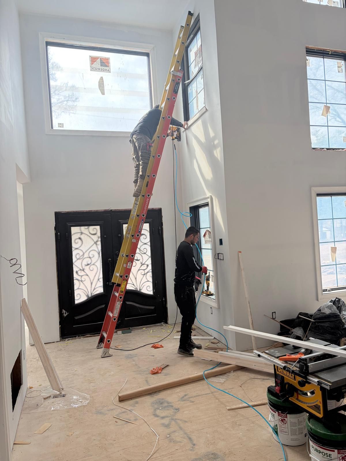 Worker on a ladder in a tall, unfinished room with large windows and construction materials scattered on the floor.