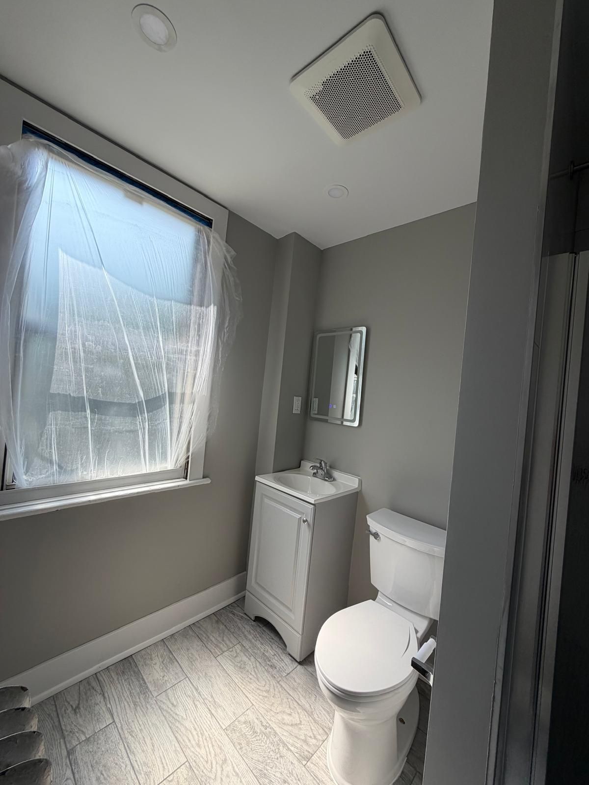 Small bathroom with a toilet, sink cabinet, mirror, and window covered with plastic sheeting.