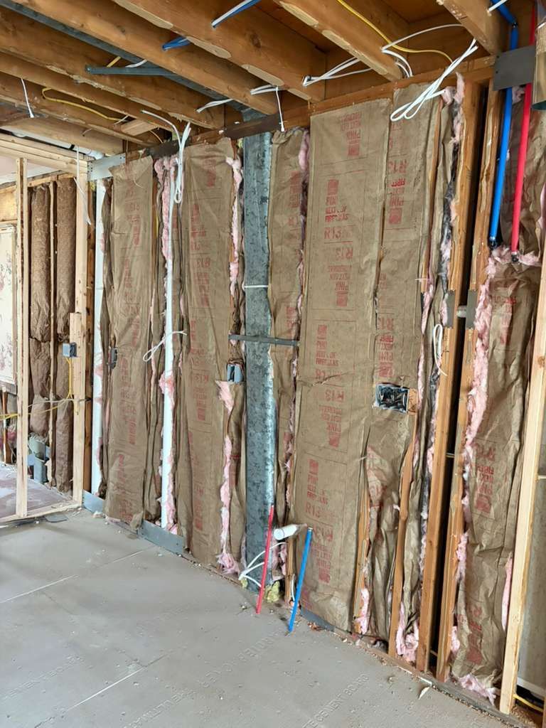 Unfinished interior wall with wooden studs, installed insulation batts, and exposed electrical wiring and plumbing pipes.
