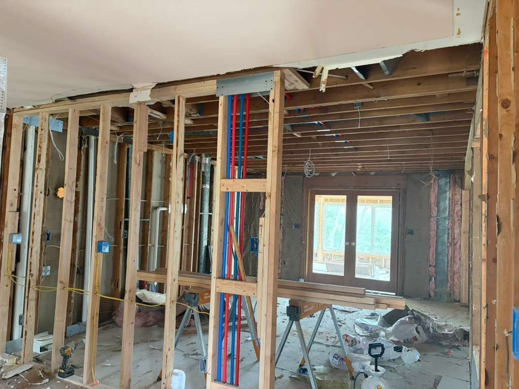 Interior view of home renovation featuring exposed wood wall studs, blue and red plumbing pipes, and a view to the outside.