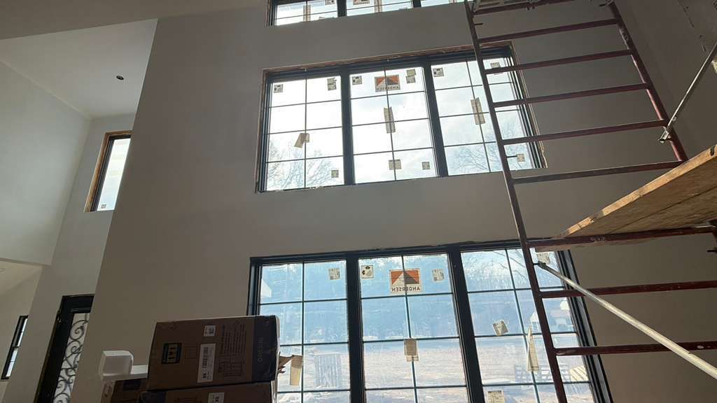 Interior view of a tall living room under construction, featuring large paneled windows and a metal scaffolding frame.
