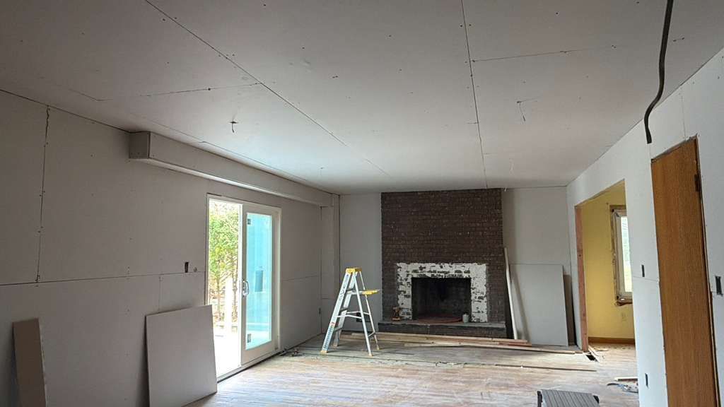 A room undergoing renovation with exposed drywall on the ceiling and walls, a brick fireplace, and a stepladder.