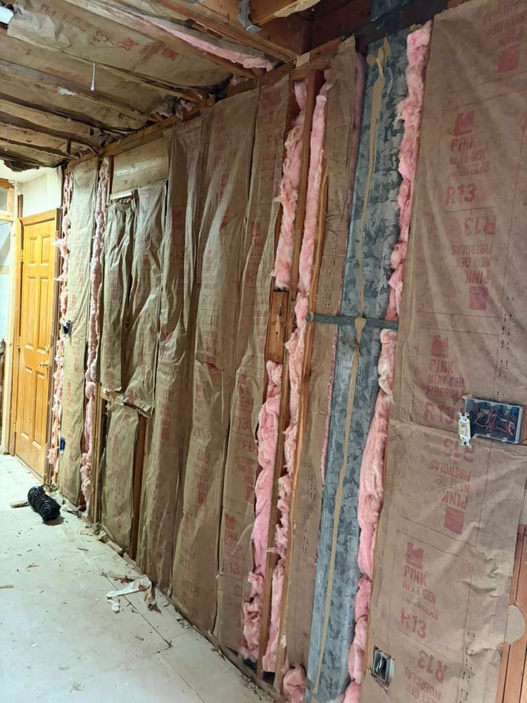 An unfinished interior wall showing exposed wooden studs, pink fiberglass insulation, and electrical outlets.