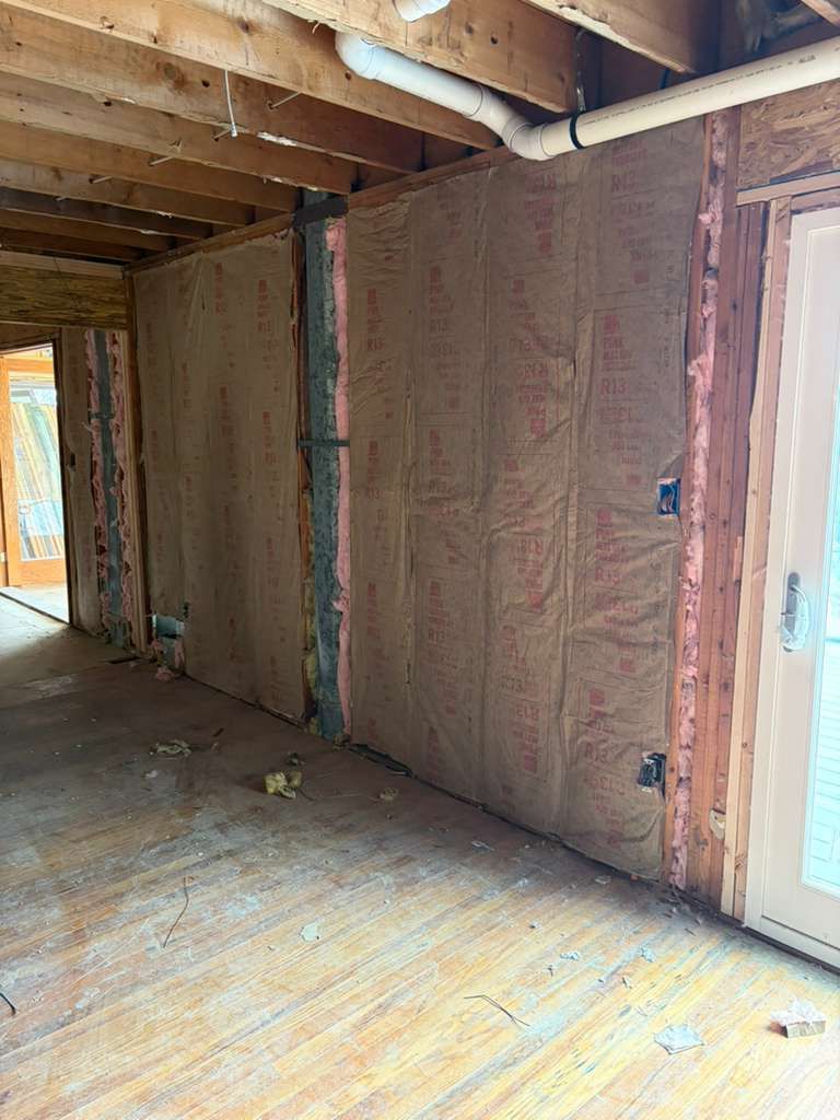 An unfinished interior room with exposed wooden ceiling joists, insulation-filled walls, and a new glass-paneled door.