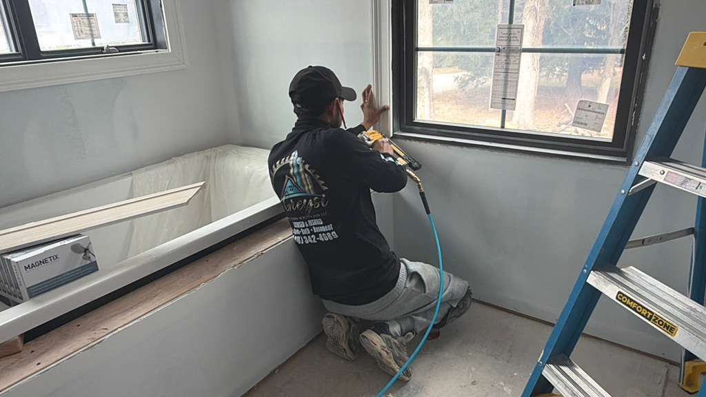 A construction worker kneeling by a bathtub, using a nail gun to install trim around a window in a room under renovation.