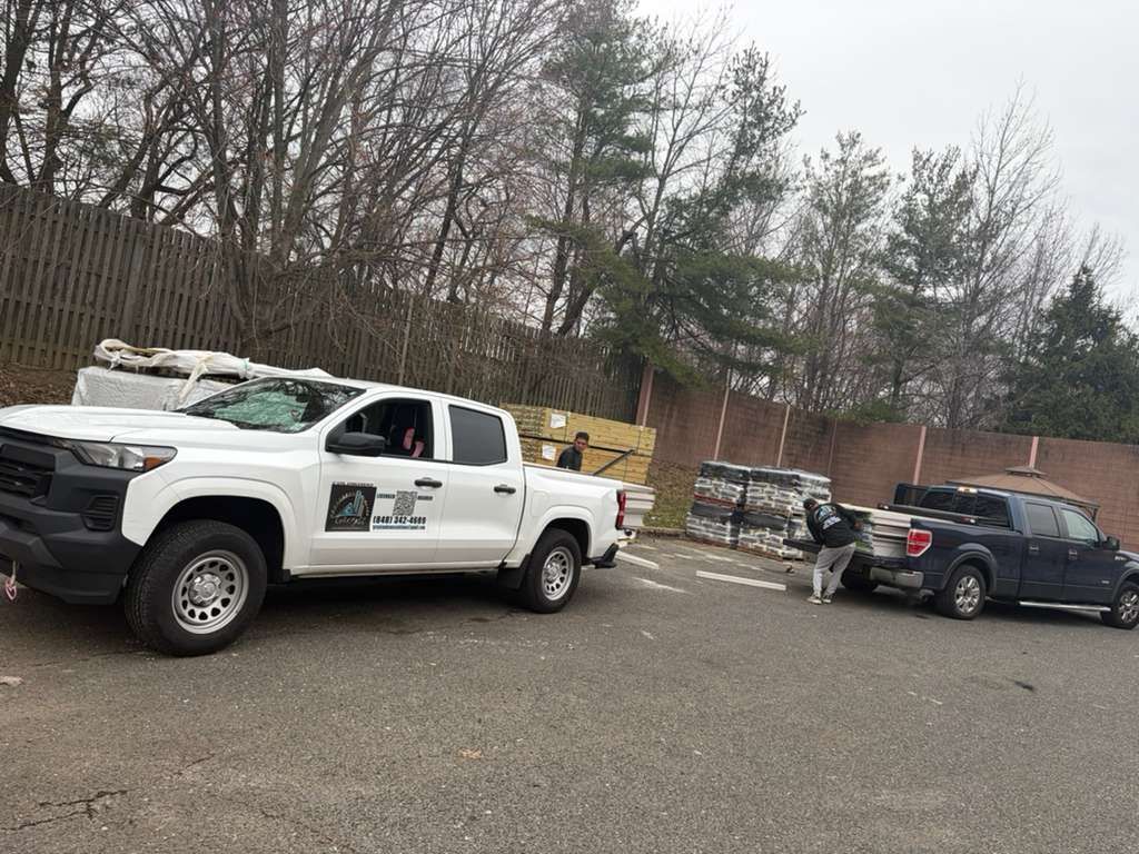 A white pickup truck and a dark blue truck parked in a gravel lot next to a wall, with workers handling cargo.