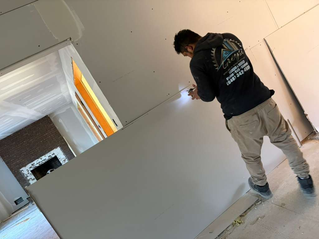 A person in a black hoodie and tan pants works on installing drywall in a room under construction with a brick fireplace.