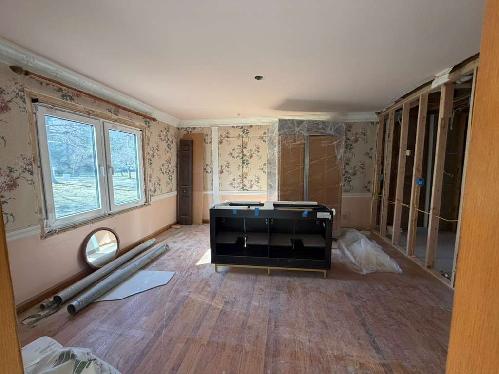 A room under renovation with wooden flooring, floral wallpaper, a center vanity cabinet, and unfinished wall framing.