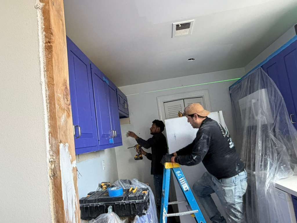 Two workers installing blue kitchen cabinets, one on a ladder using a power drill in a room undergoing renovation.