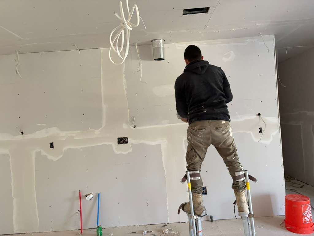 A construction worker on stilts applies joint compound to drywall seams on a wall in an unfinished room.