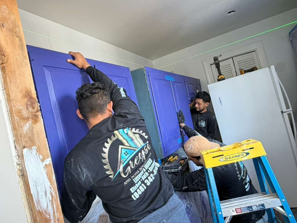 Two workers in black shirts install purple cabinets in a room with white walls and a green laser level line.