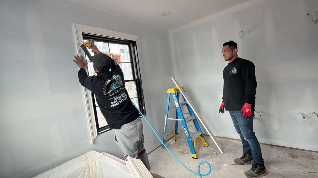 Two people installing trim around a window in a room with light-gray drywall, a step ladder, and a blue air hose.