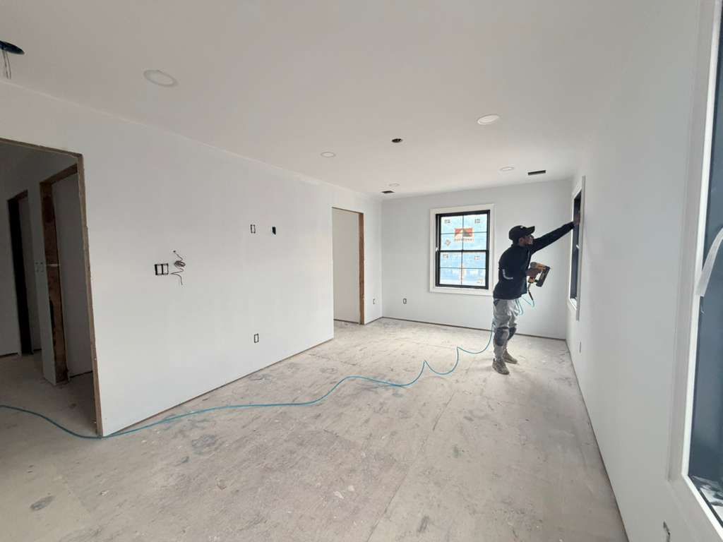 A construction worker finishes drywall around a window in an unfinished, white-walled room with a concrete subfloor.