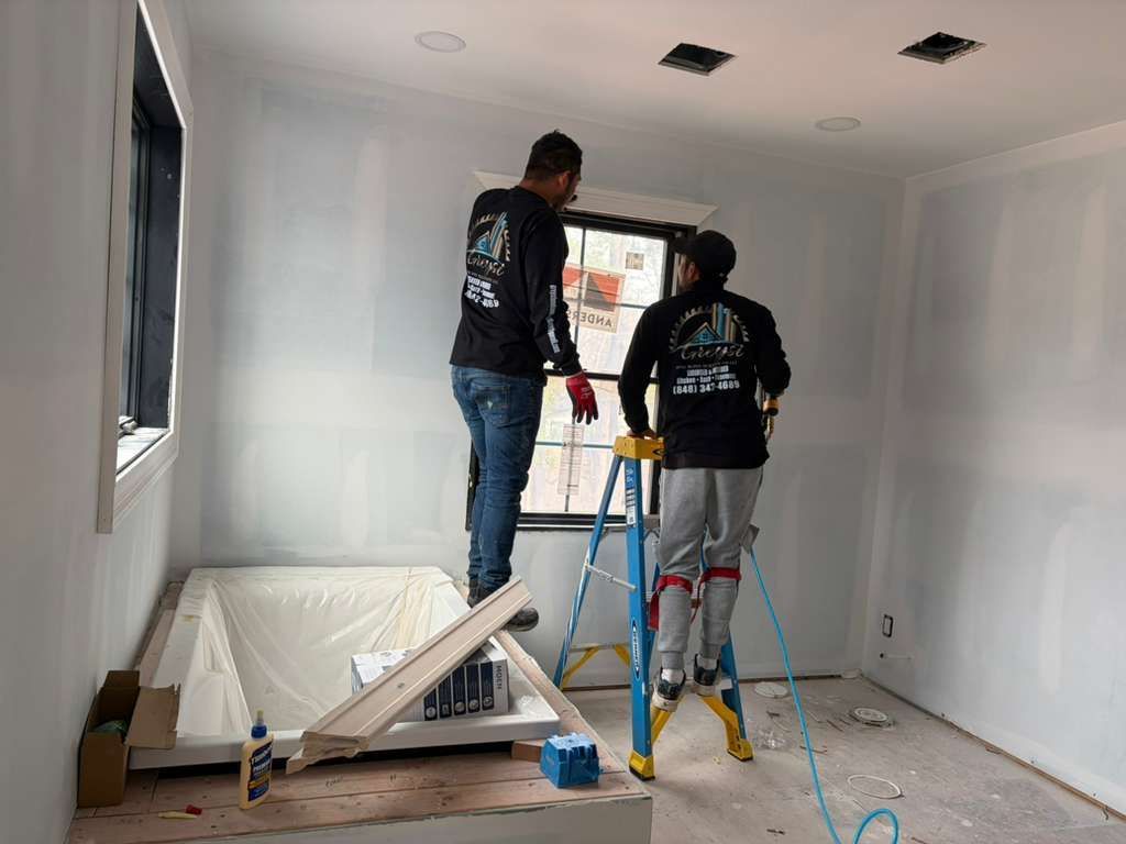 Two workers in black uniforms install a window frame in a room with unfinished drywall and a built-in bathtub.