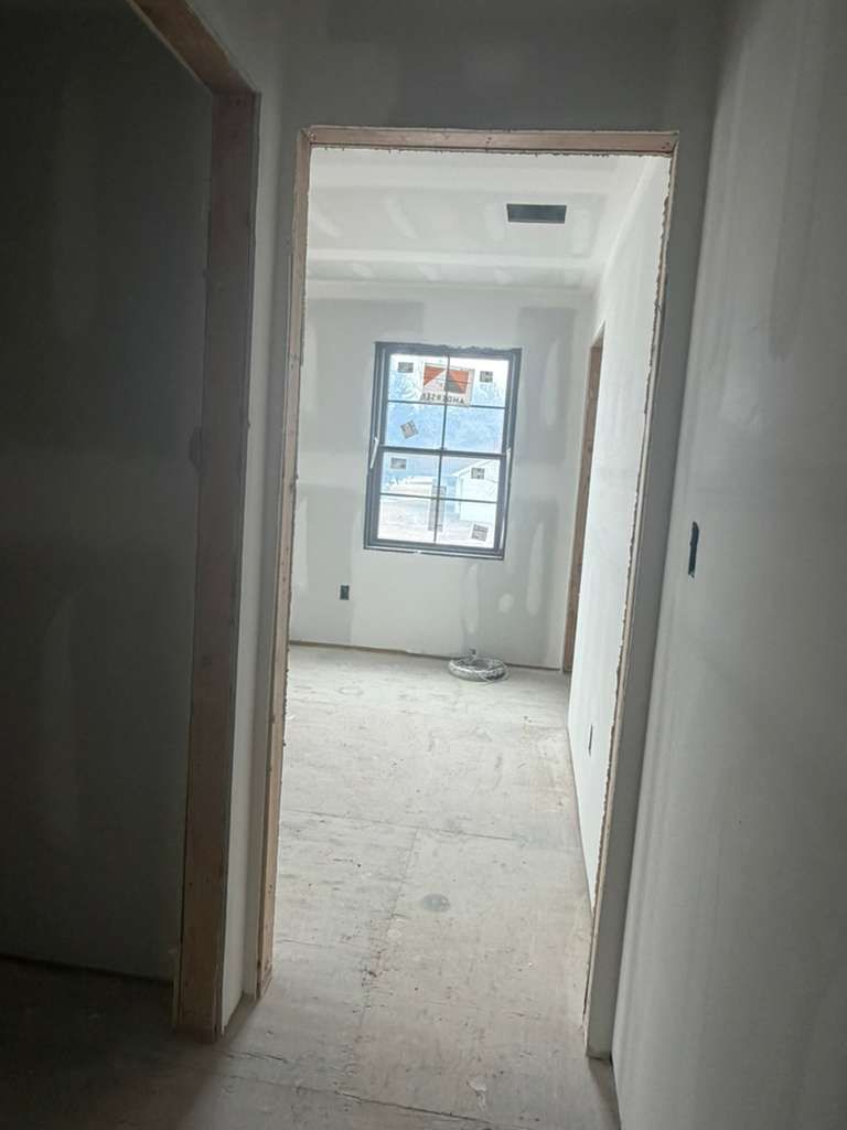 An interior view of a hallway leading to a room under construction with bare drywall, wood frames, and a visible window.