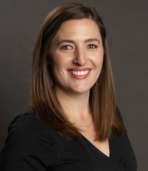 A person with shoulder-length brown hair wearing a black top, smiling against a dark gray background.