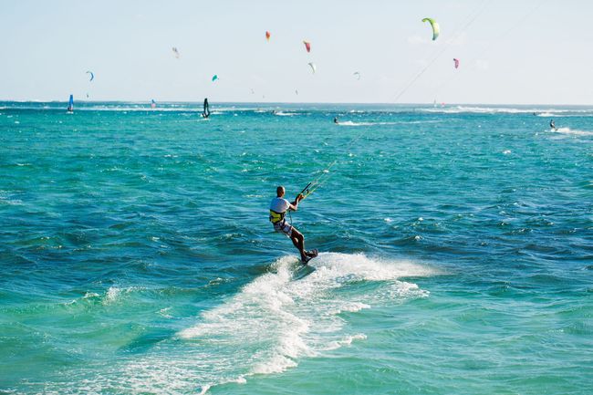 Kitesurfer gliding across turquoise ocean waves with colorful kites in the sky