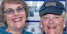 Two older women wearing glasses and a street pastor hat are smiling for the camera.