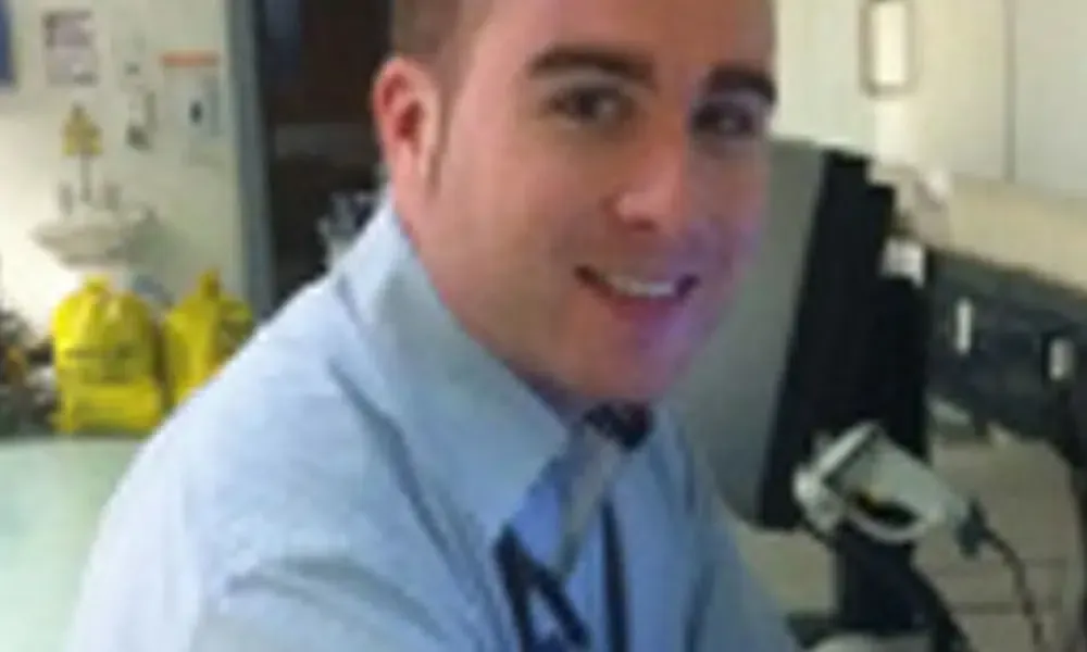 A man in a blue shirt and tie is smiling in front of a computer monitor.