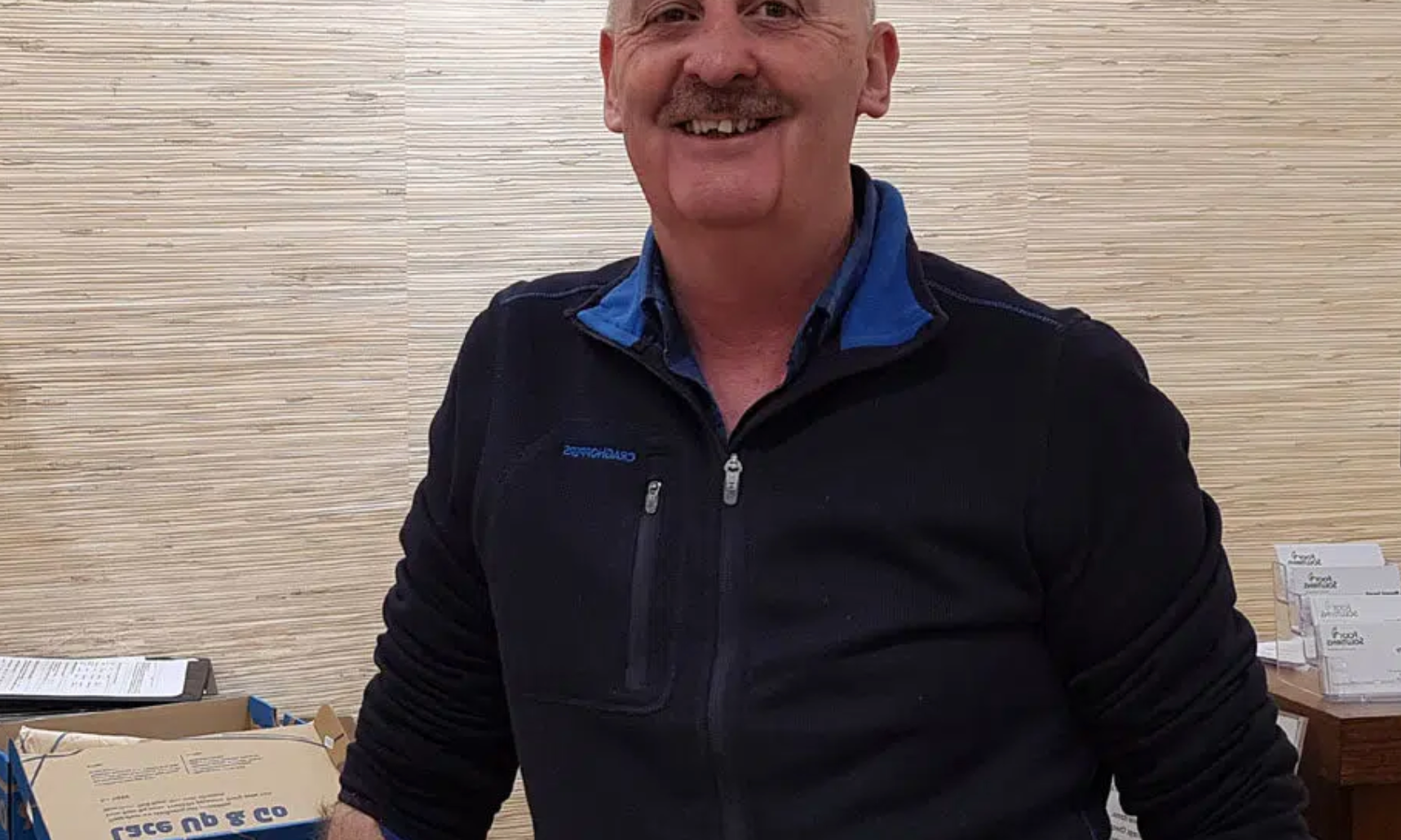 A man with a mustache is smiling for the camera while standing in front of a wooden wall.