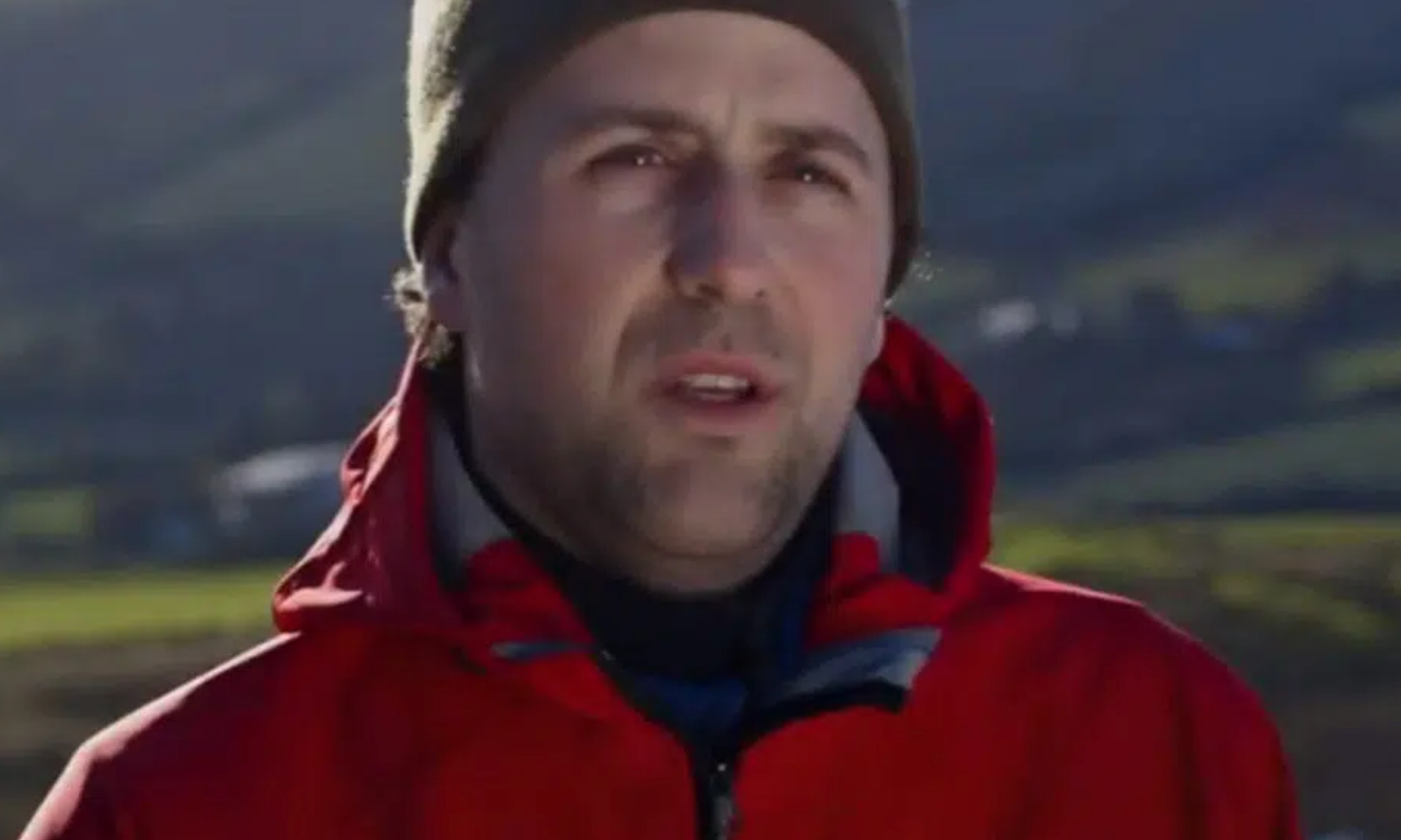 A man wearing a red jacket and a hat is standing in front of a mountain.