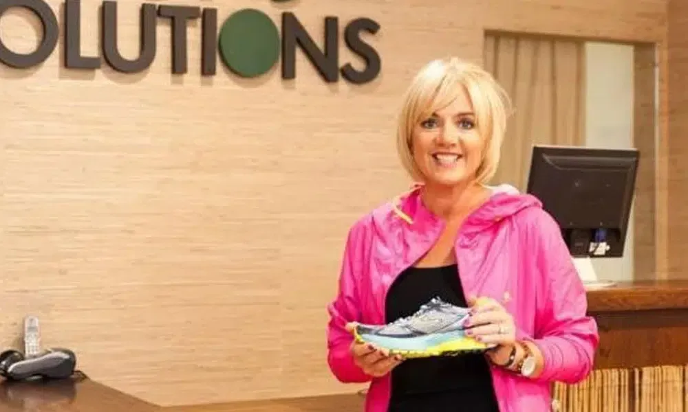 A woman in a pink jacket is holding a pair of shoes in front of a sign that says solutions.