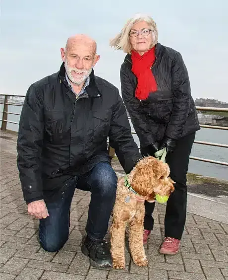 A man and a woman are kneeling down next to a dog with a tennis ball in its mouth.