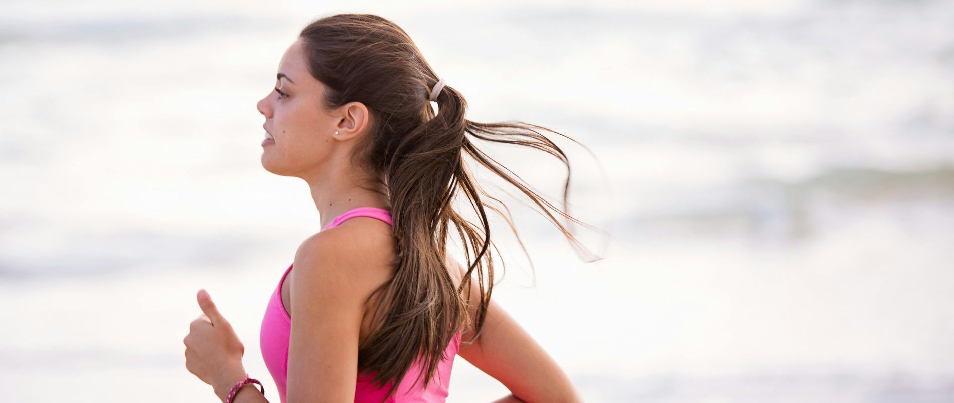 A person with a ponytail wearing a pink top runs along a beach with a blurred, bright background.