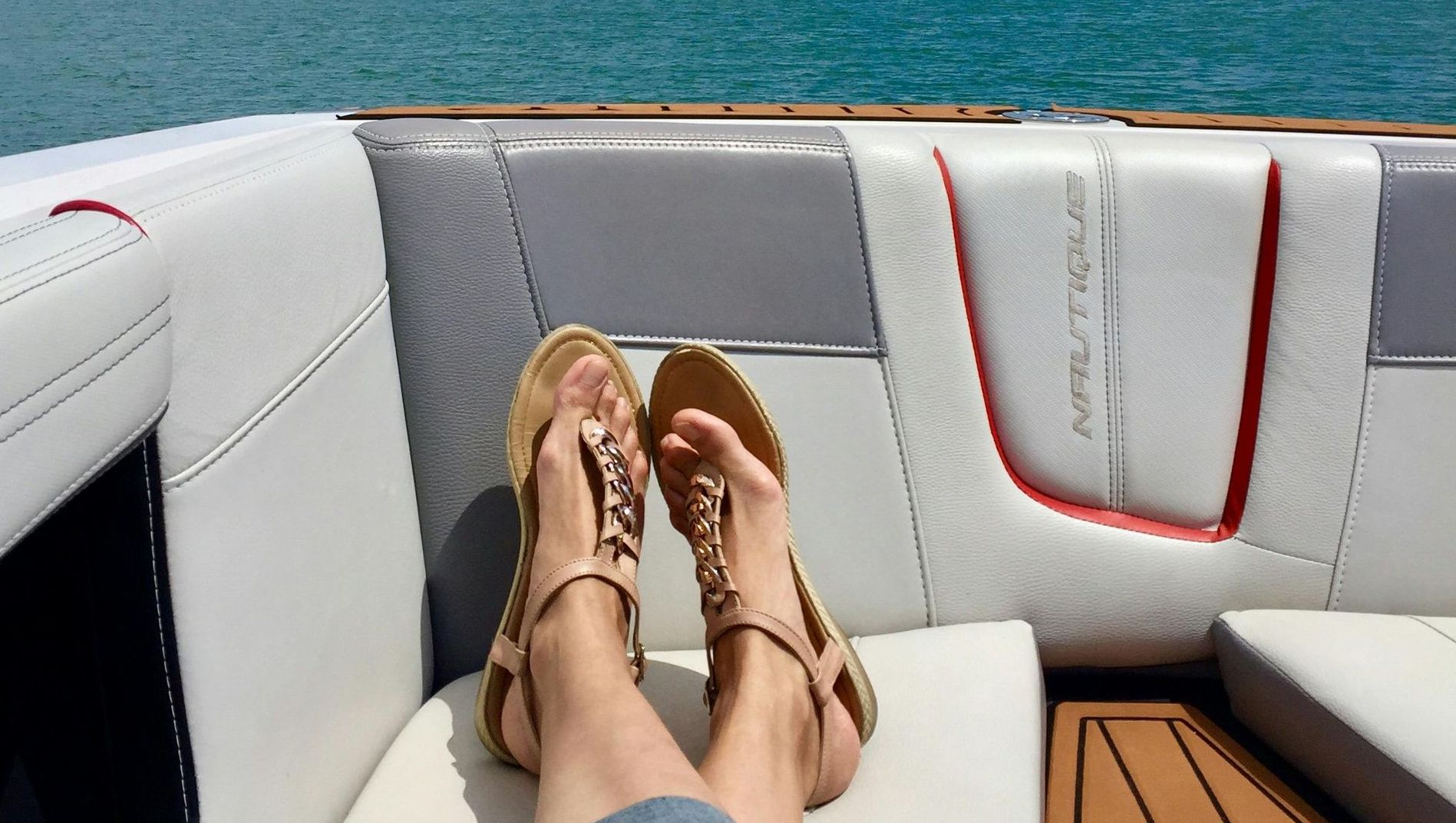 A person's feet in tan sandals resting on white boat cushions, with a view of turquoise water in the background.