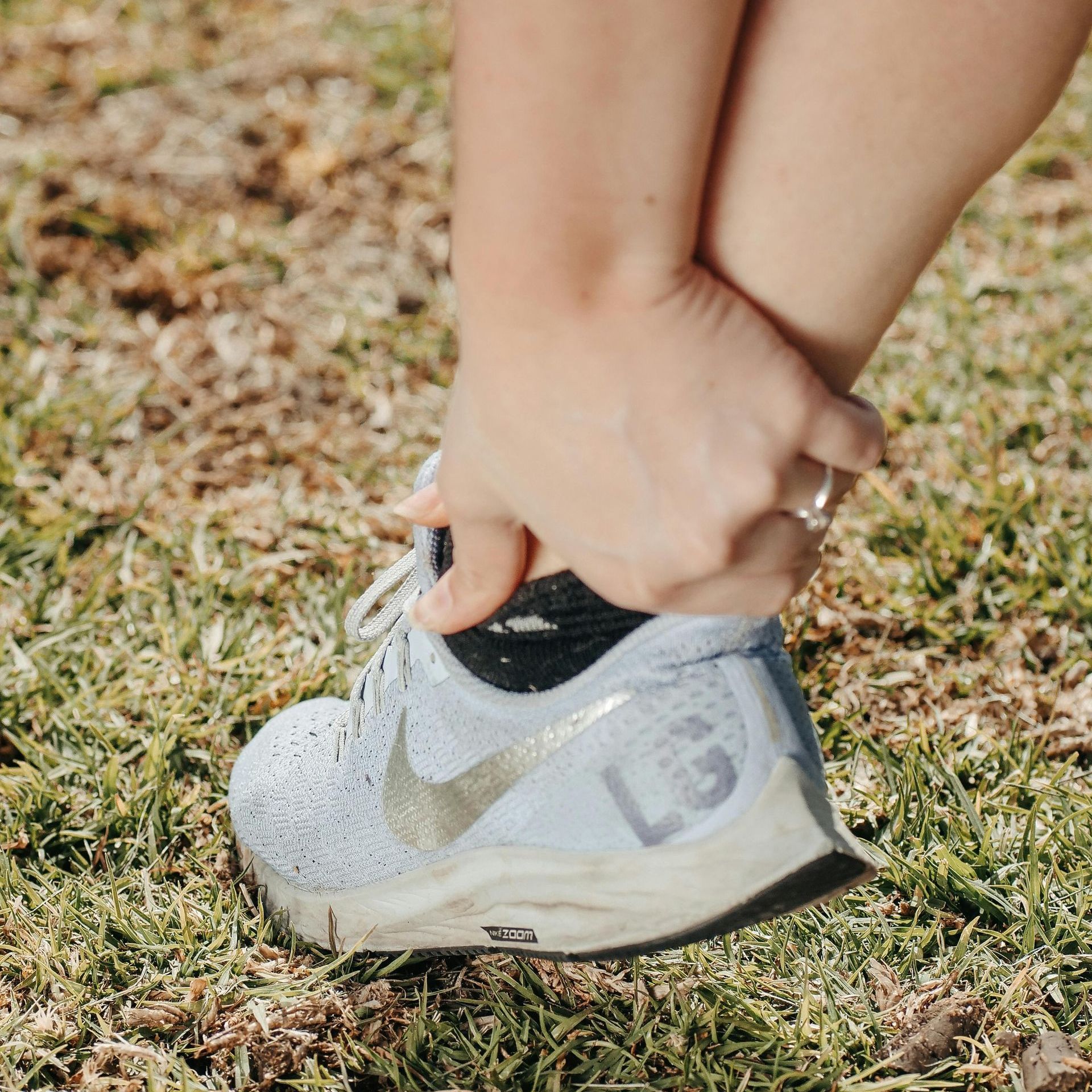 A person holds their ankle while wearing a white Nike sneaker on grass, suggesting a possible sports injury.