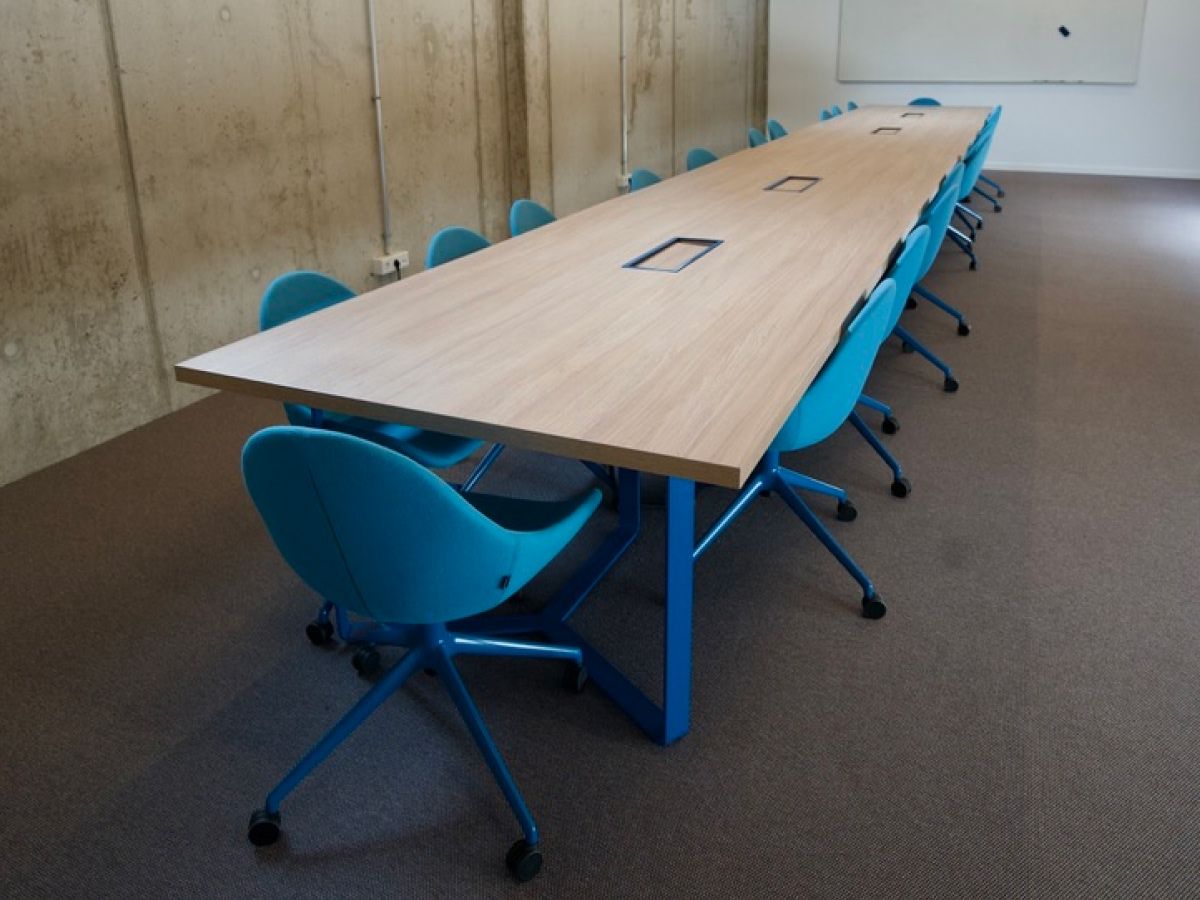Ein langer Konferenztisch mit heller Holzplatte und blauen Beinen, umgeben von passenden blauen Bürostühlen auf einem Teppichboden.