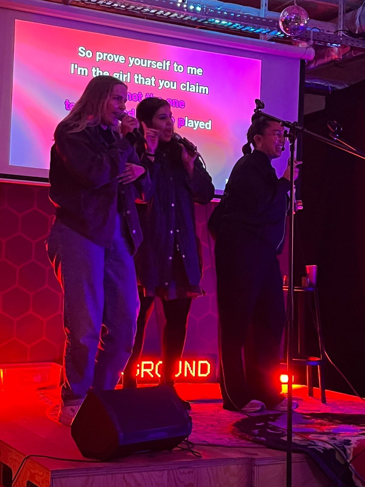 Drei Personen singen Karaoke auf einer Bühne mit einer hell erleuchteten Leinwand im Hintergrund, die in rotes Bühnenlicht getaucht ist.