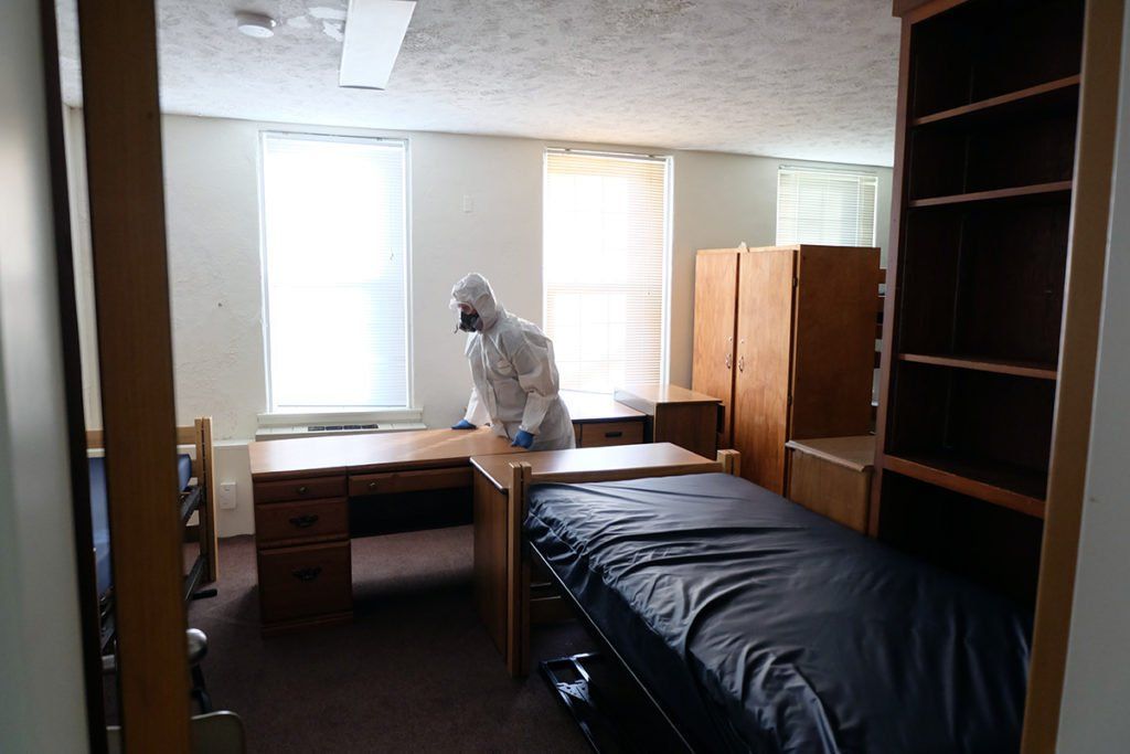 A man in a protective suit is cleaning a dorm room.