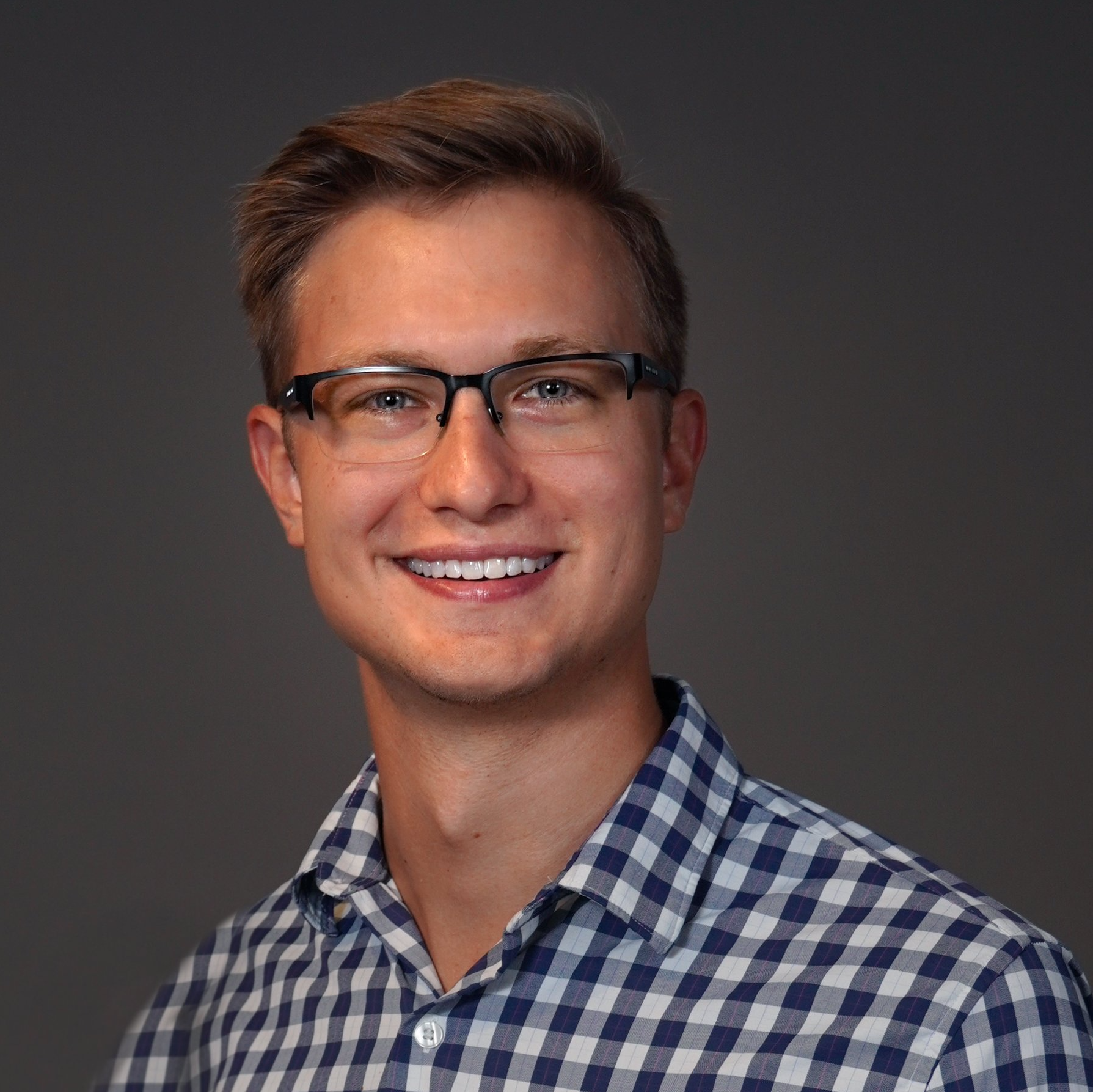 A young man wearing glasses and a plaid shirt is smiling for the camera.
