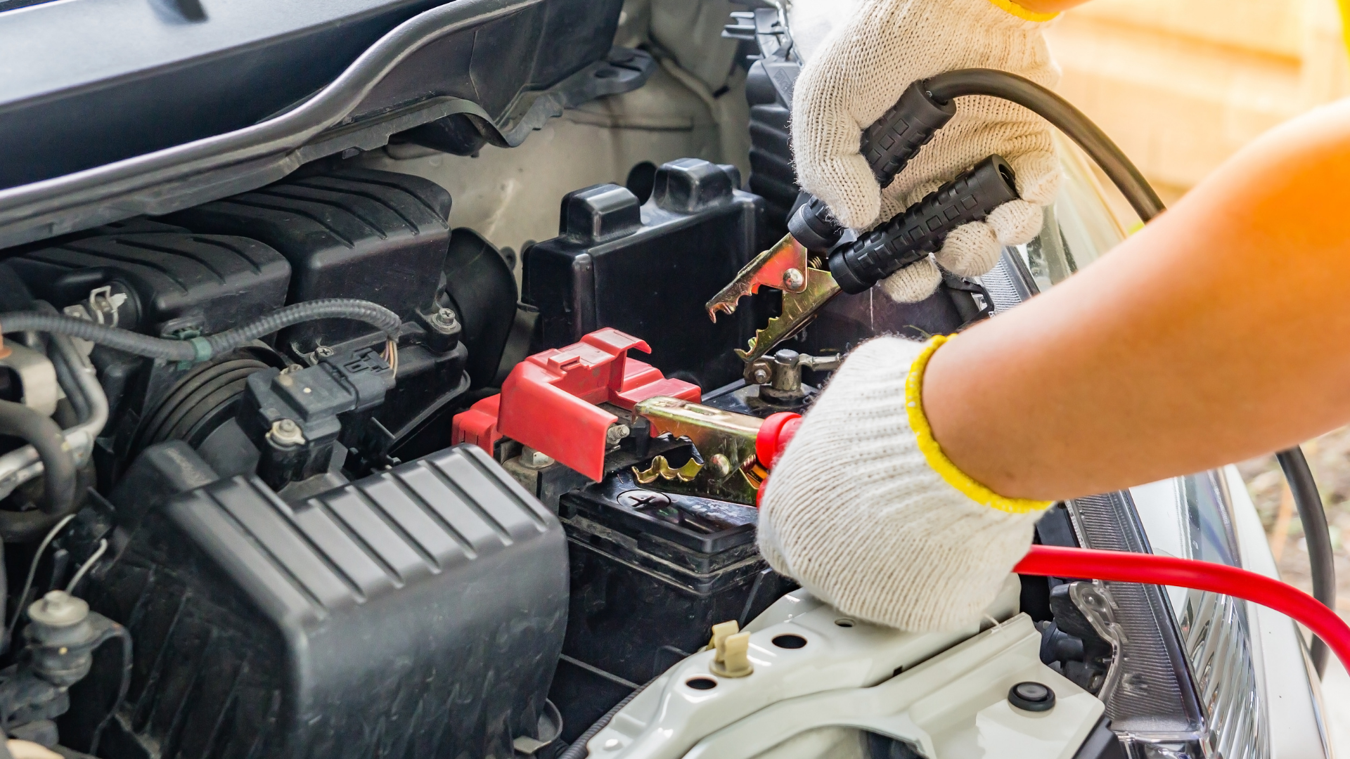 Mechanic’s gloved hands working on a car battery under the hood with red jumper cables