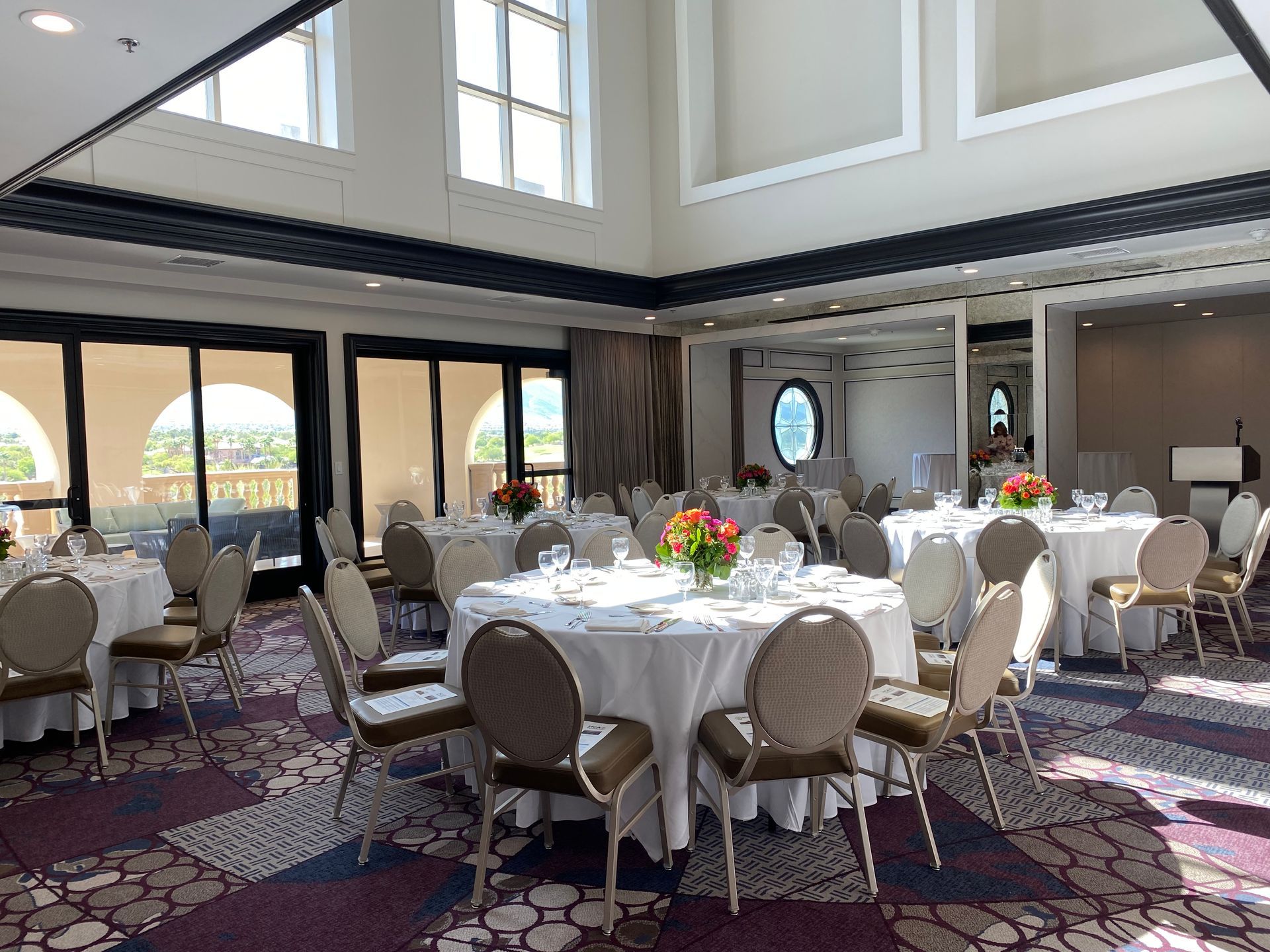 Elegant banquet hall with round tables set for a meal. Sunlight streams in through large windows; red and orange flowers are centerpieces.