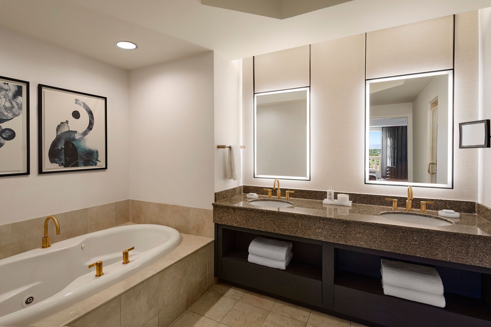 A modern bathroom with a bathtub and a double vanity with gold fixtures. Two framed mirrors and artwork decorate the white walls.