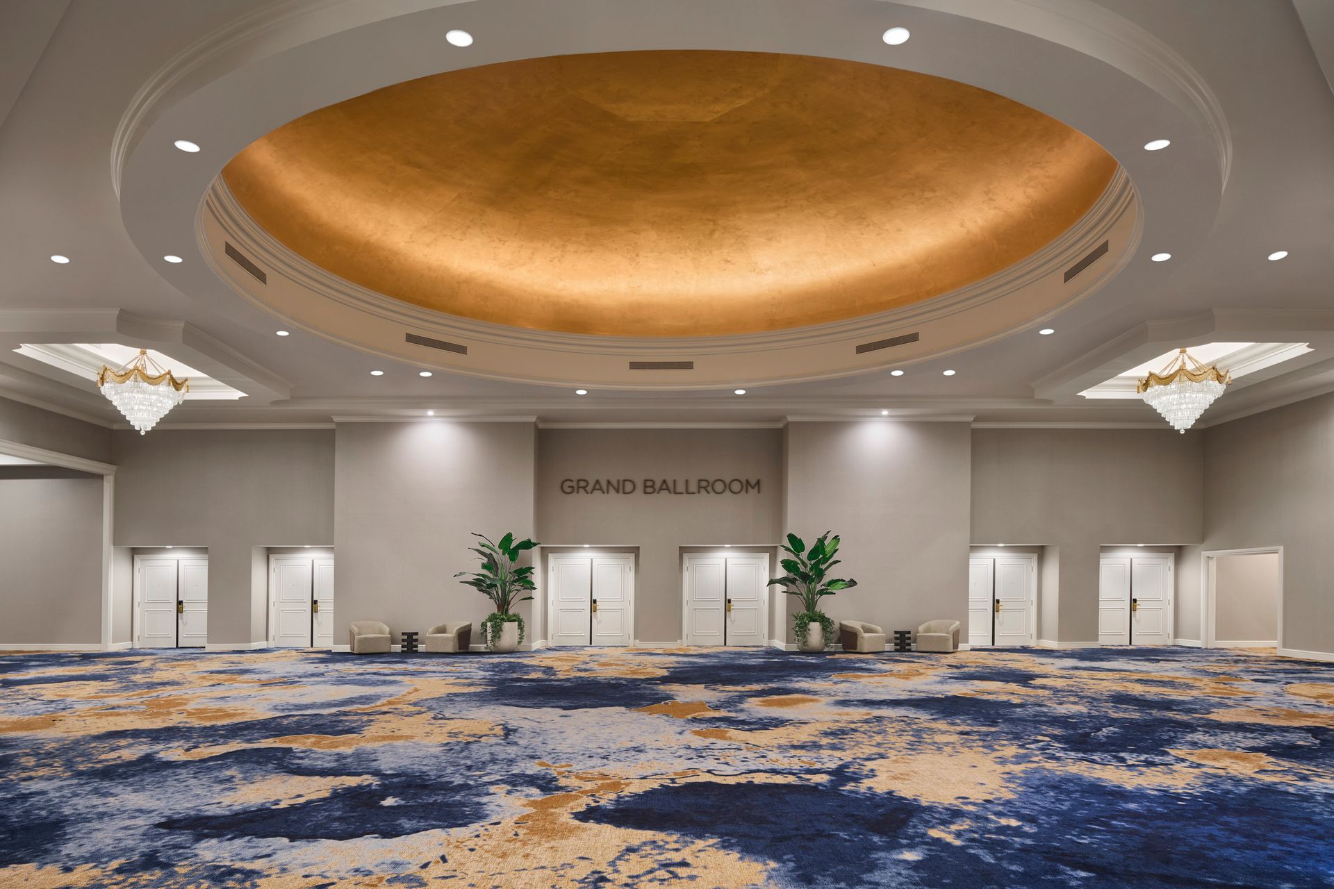 Grand ballroom with a patterned blue and gold carpet, neutral walls, and a gold-domed ceiling. Multiple doors line the back wall.