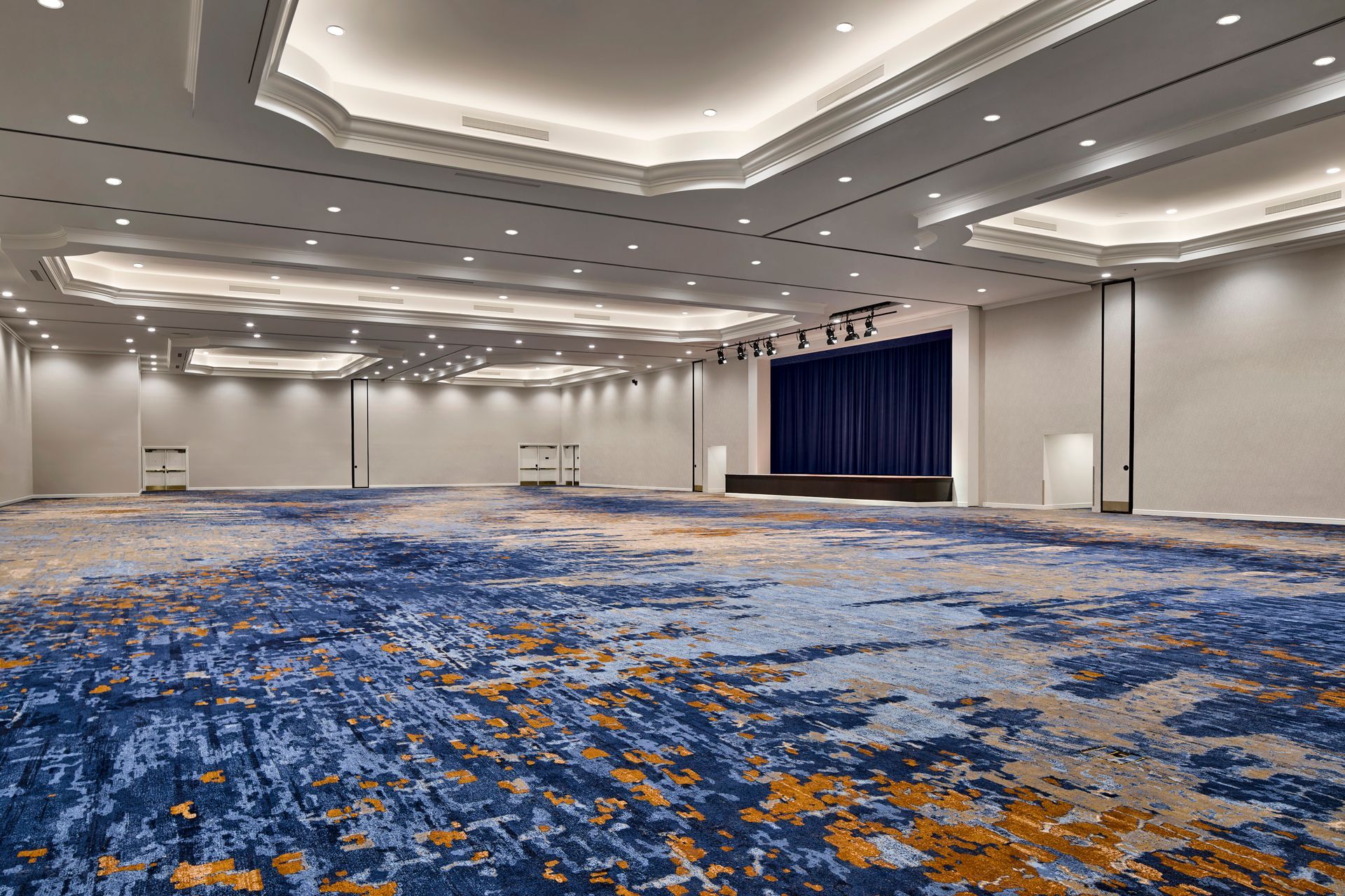 Large, empty event space with blue, gold, and white patterned carpet, a stage with blue curtains, and recessed ceiling lights.