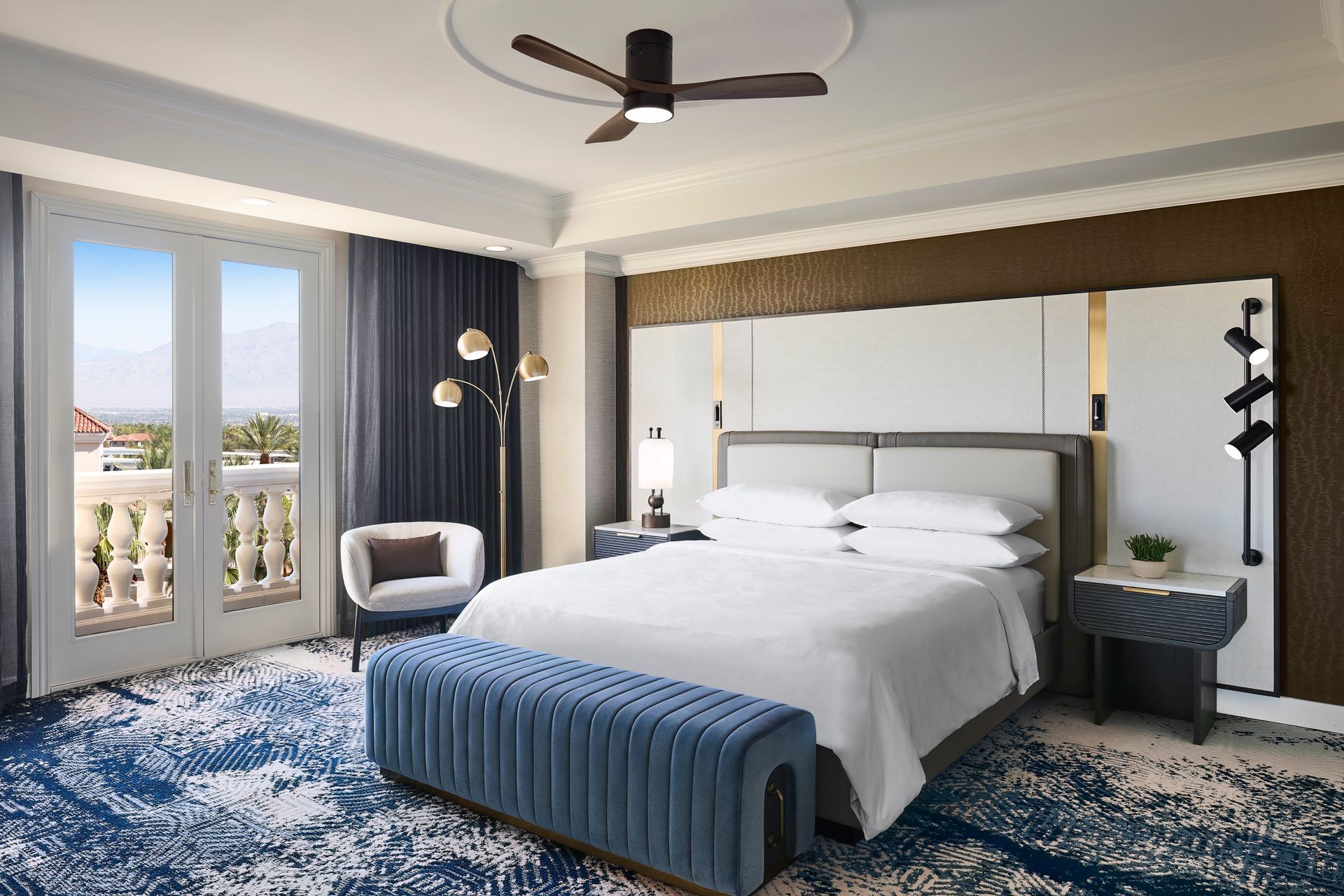 Elegant hotel bedroom with a king-sized bed, balcony doors, blue rug, and a ceiling fan.