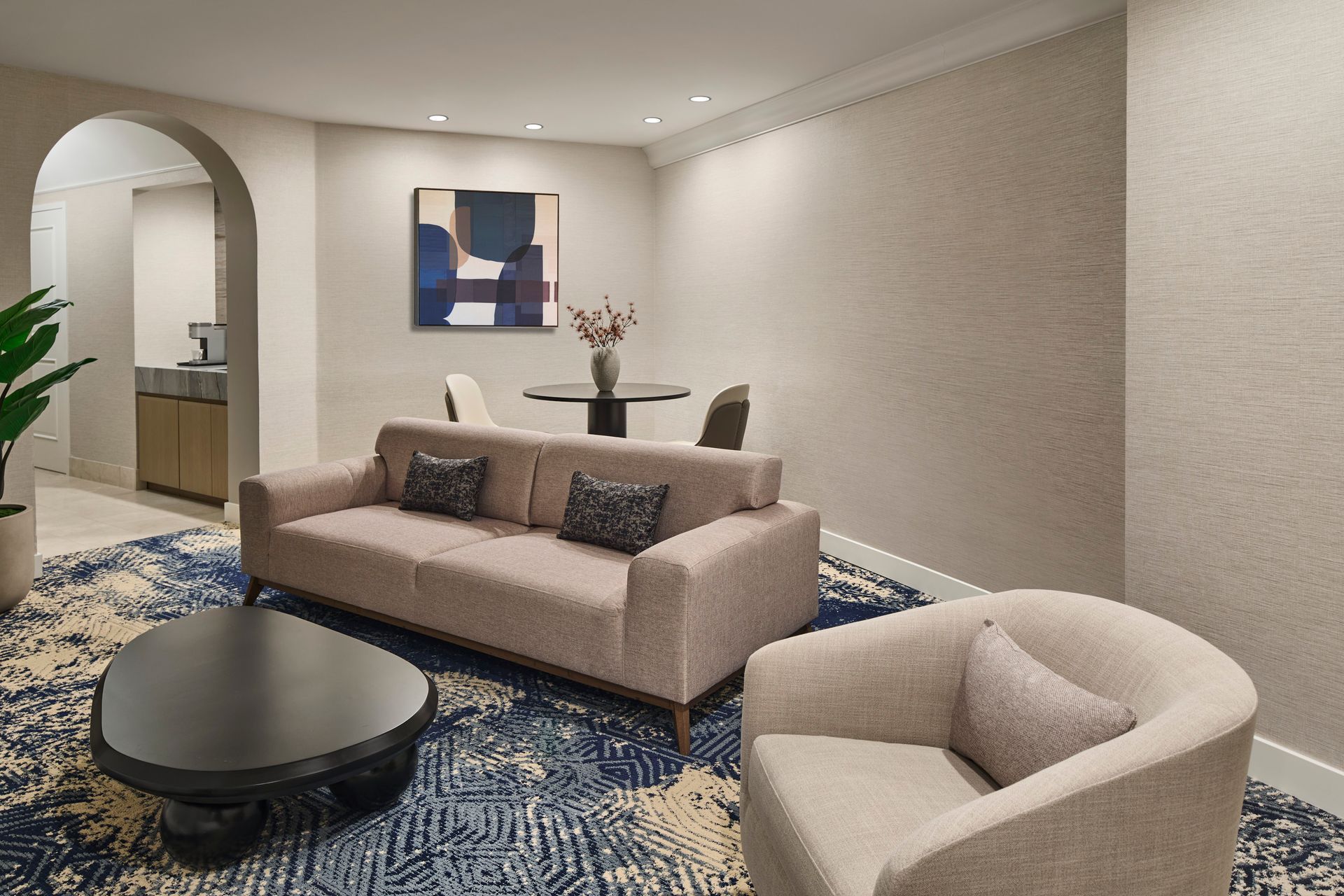 Cozy hotel living space with beige sofa, chair, and dark coffee table on a patterned rug. Round dining table and archway visible in the background.