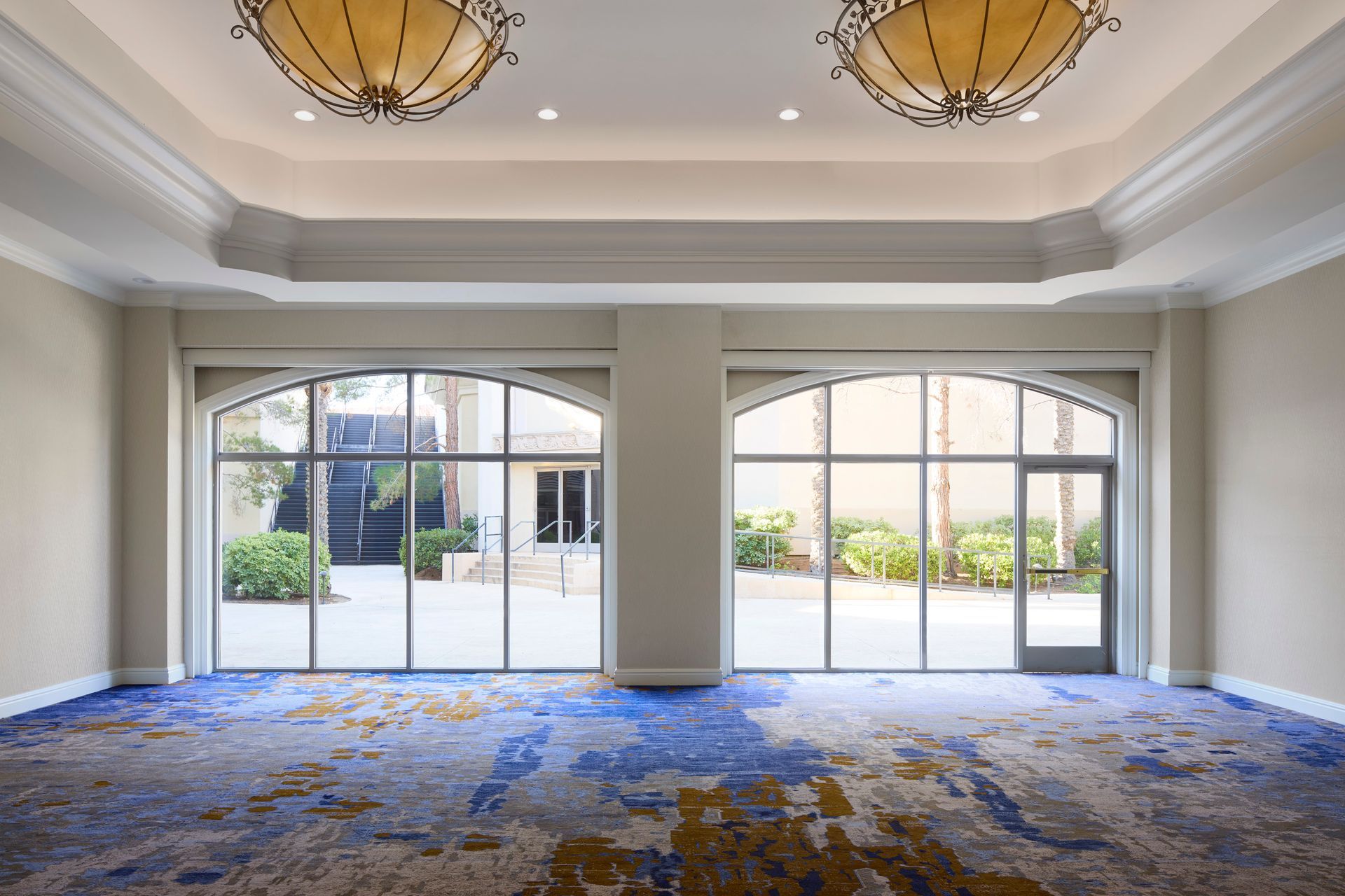 An empty room with blue and gold patterned carpet and large arched windows overlooking a courtyard. Two ornate light fixtures hang from the ceiling.