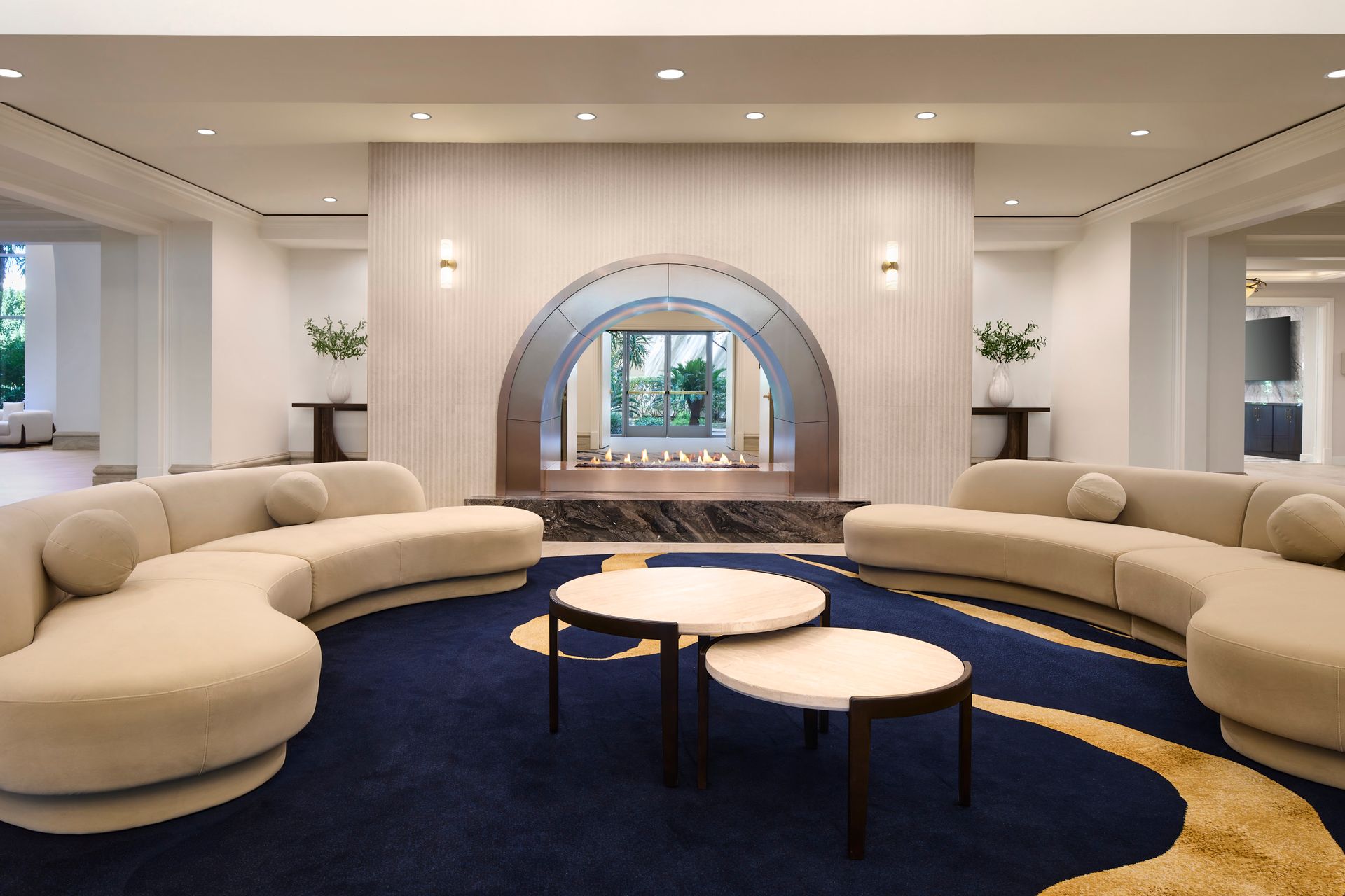 Spacious, modern hotel lobby with curved cream-colored couches, round coffee tables, and a dark blue rug with gold accents, centered around an arched feature.