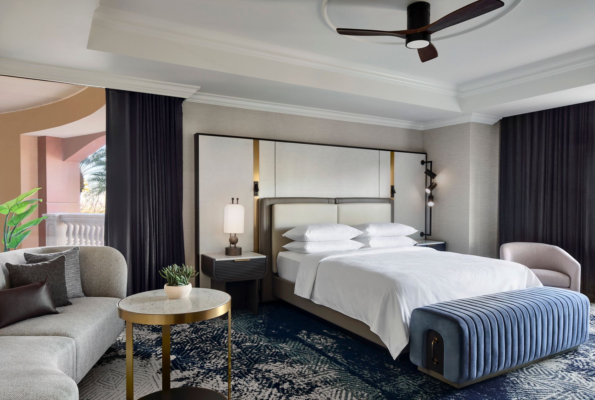 A hotel bedroom with a large bed, sofa, and balcony access. Features include dark blue curtains, a patterned rug, and a blue velvet ottoman.