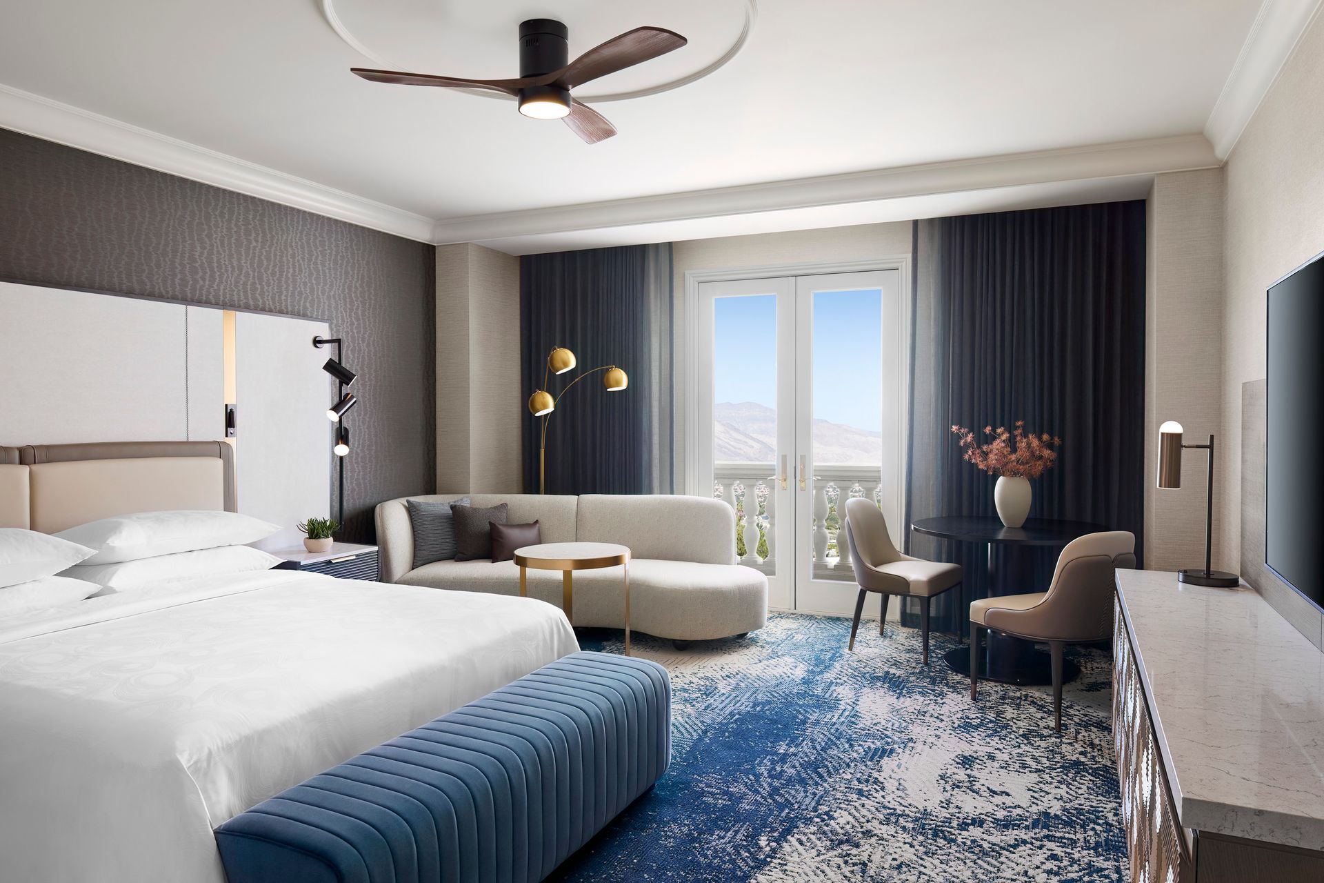 A luxurious hotel room with a large bed, sofa, and balcony, featuring blue and neutral tones.