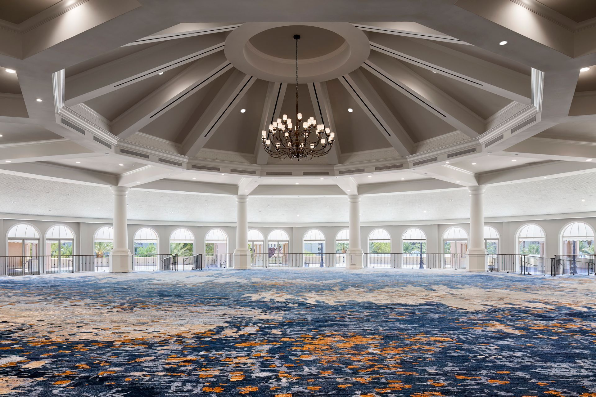 An expansive, circular ballroom with a patterned blue and gold carpet and a central chandelier beneath a domed ceiling.