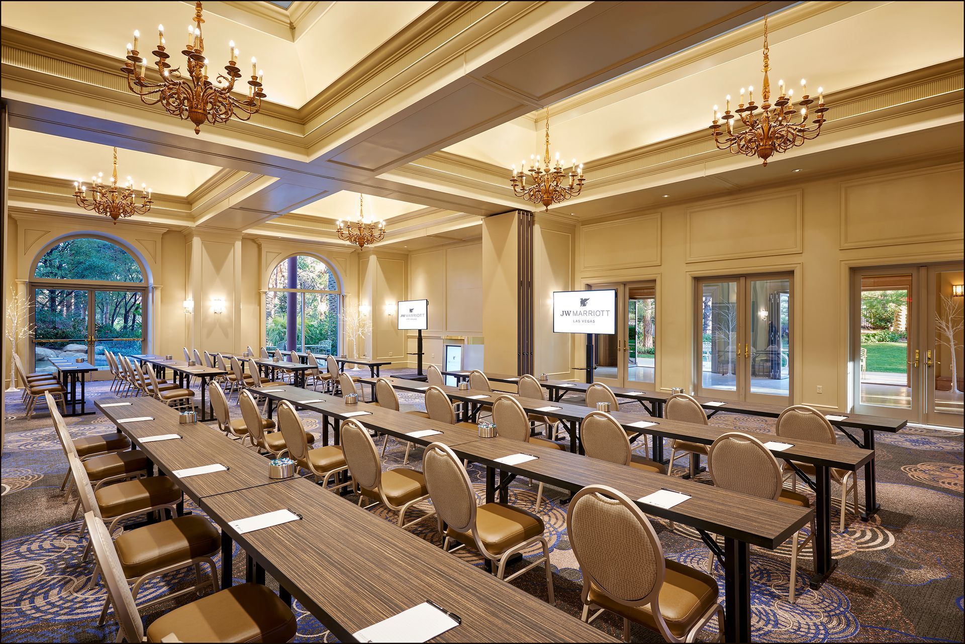 A formal meeting room with rows of tables and chairs set up for a presentation. Large windows and chandeliers provide light.