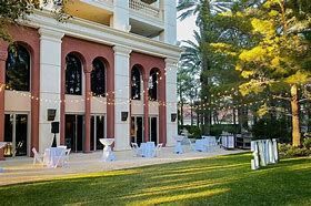 Outdoor event setup with tables, chairs, and string lights in front of a building with large arched windows. Green lawn, trees.